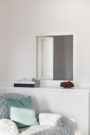 Ein Zimmer mit weißer Wand, einem Fenster mit weißem Rahmen, einem Heizkörper, einem Bücherstapel und einem Kissen auf einem Stuhl.