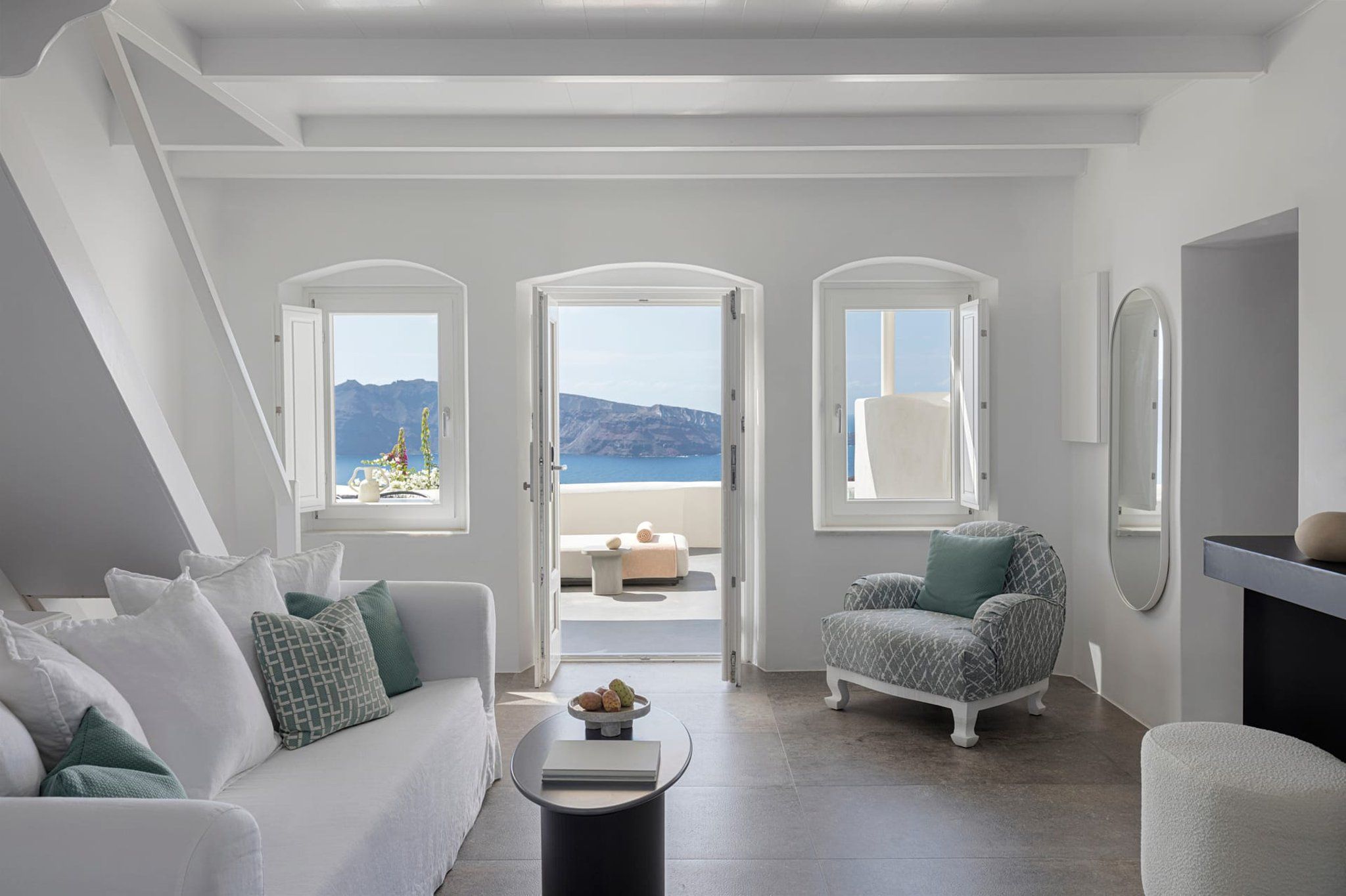 Un salon moderne avec un canapé blanc, une table basse ronde et un fauteuil à motifs. Trois fenêtres en arc offrent une vue sur la mer et les montagnes.
