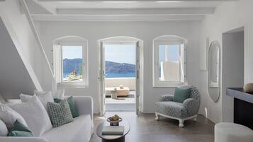Un salon moderne avec un canapé blanc, une table basse ronde et un fauteuil à motifs. Trois fenêtres en arc offrent une vue sur la mer et les montagnes.