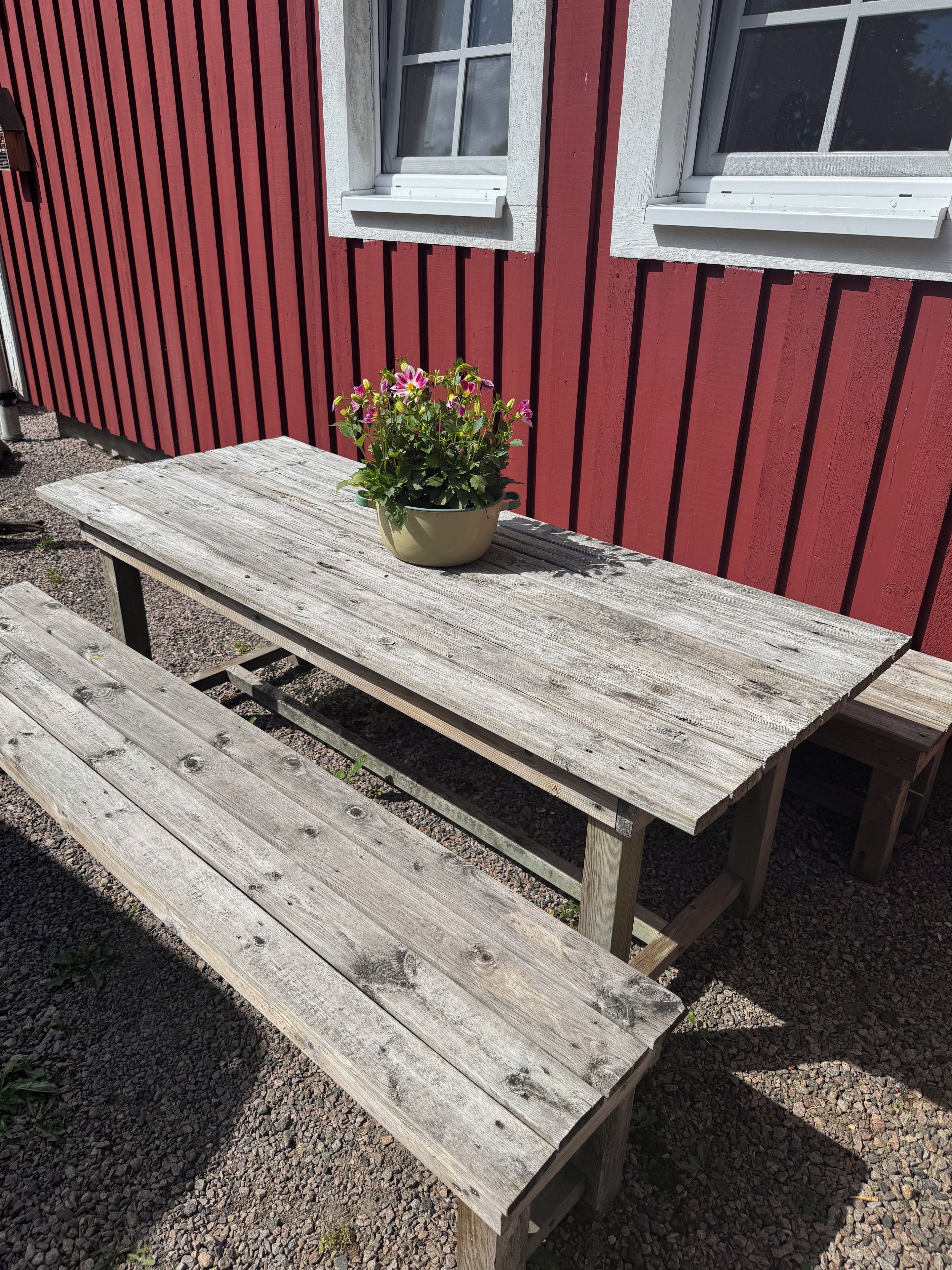 A wooden picnic table with a potted plant in front of a red building with white window frames.
