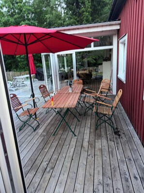 Een terras buiten met een houten tafel en stoelen, een rode parasol en een rood gebouw op de achtergrond.
