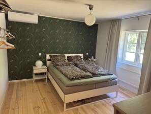 Een slaapkamer met een bed, nachtkastje, wandairconditioner, raam met gordijnen en een hanglamp.