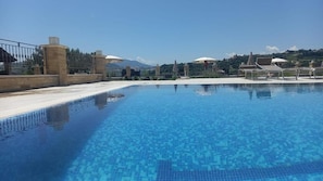 A swimming pool with a tiled floor, surrounded by lounge chairs and umbrellas, with a view of hills in the background.