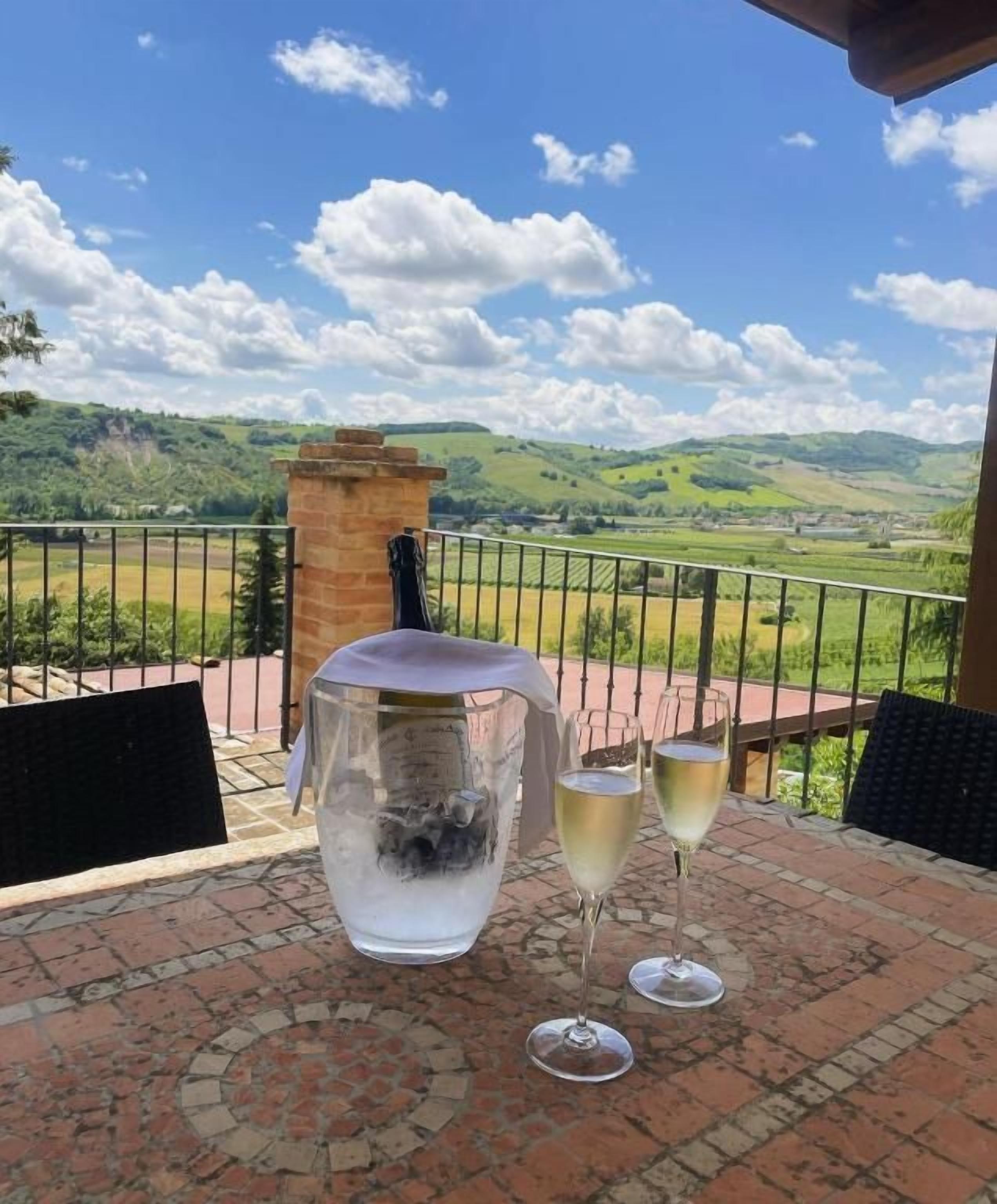 A terrace with a table, two glasses of white wine, and a large glass container.