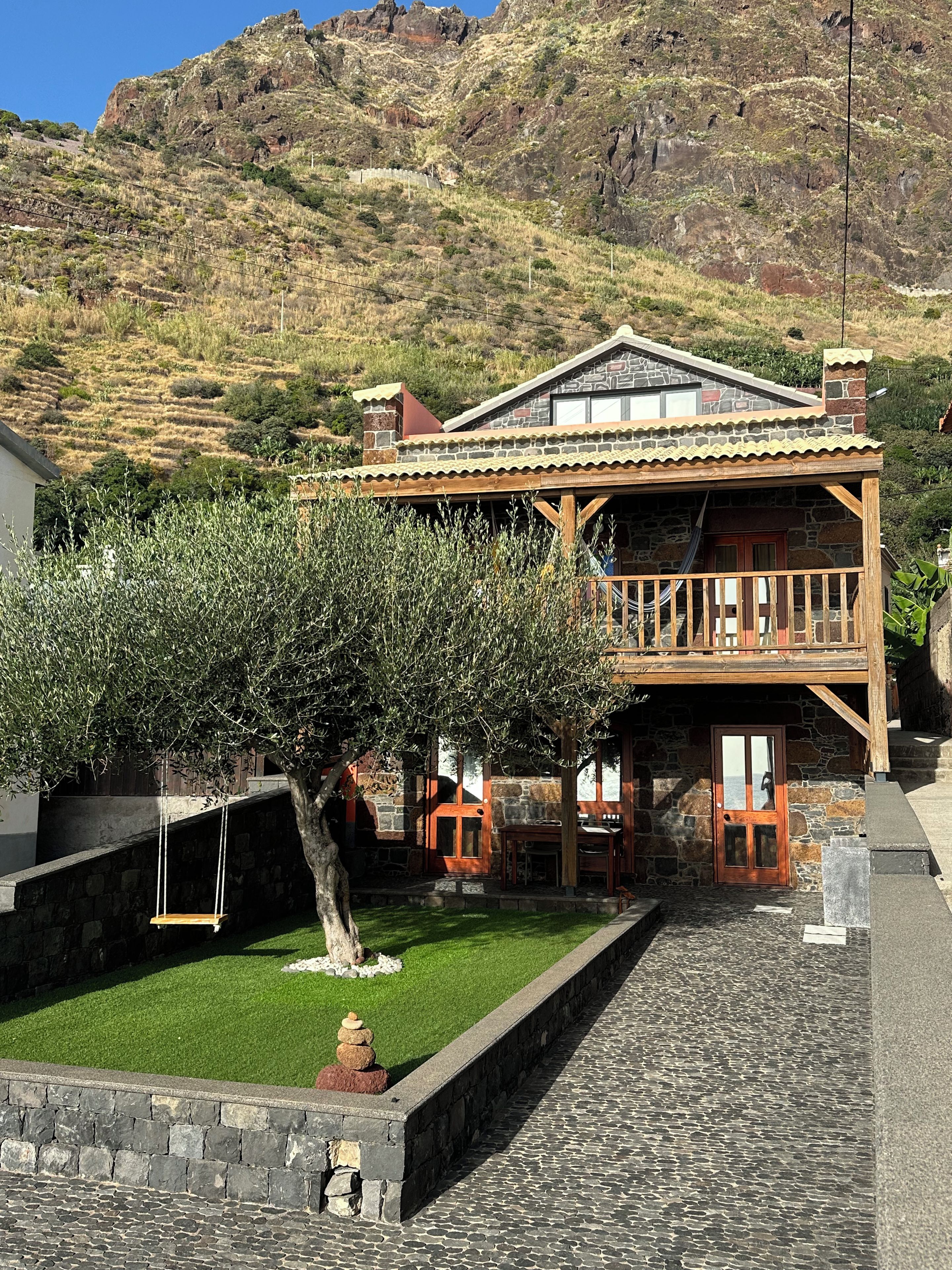 A stone building with a wooden balcony, a swing, and a small garden with a tree.