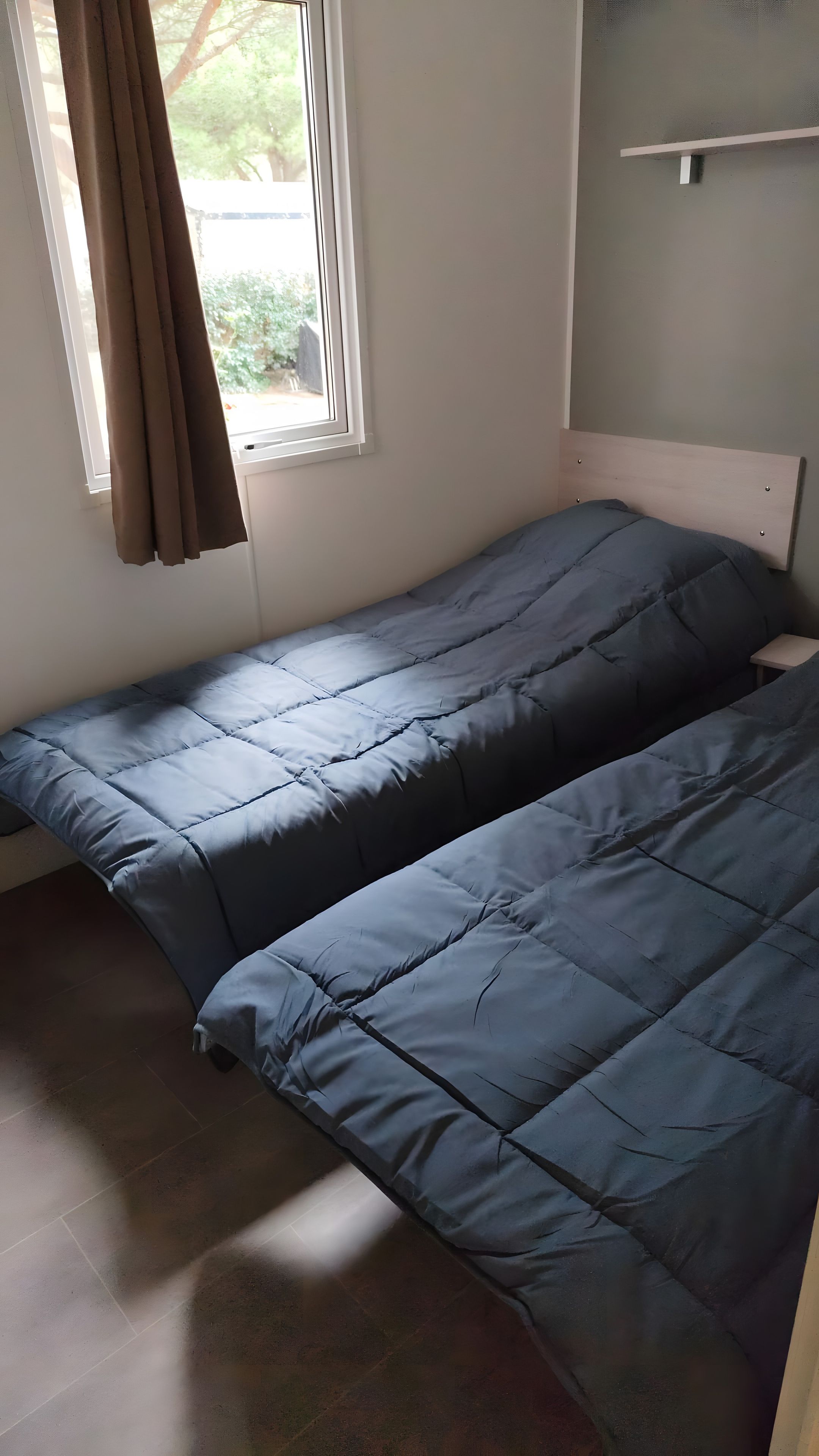Une petite chambre avec un lit simple, une fenêtre, une tête de lit en bois et une petite table de chevet.