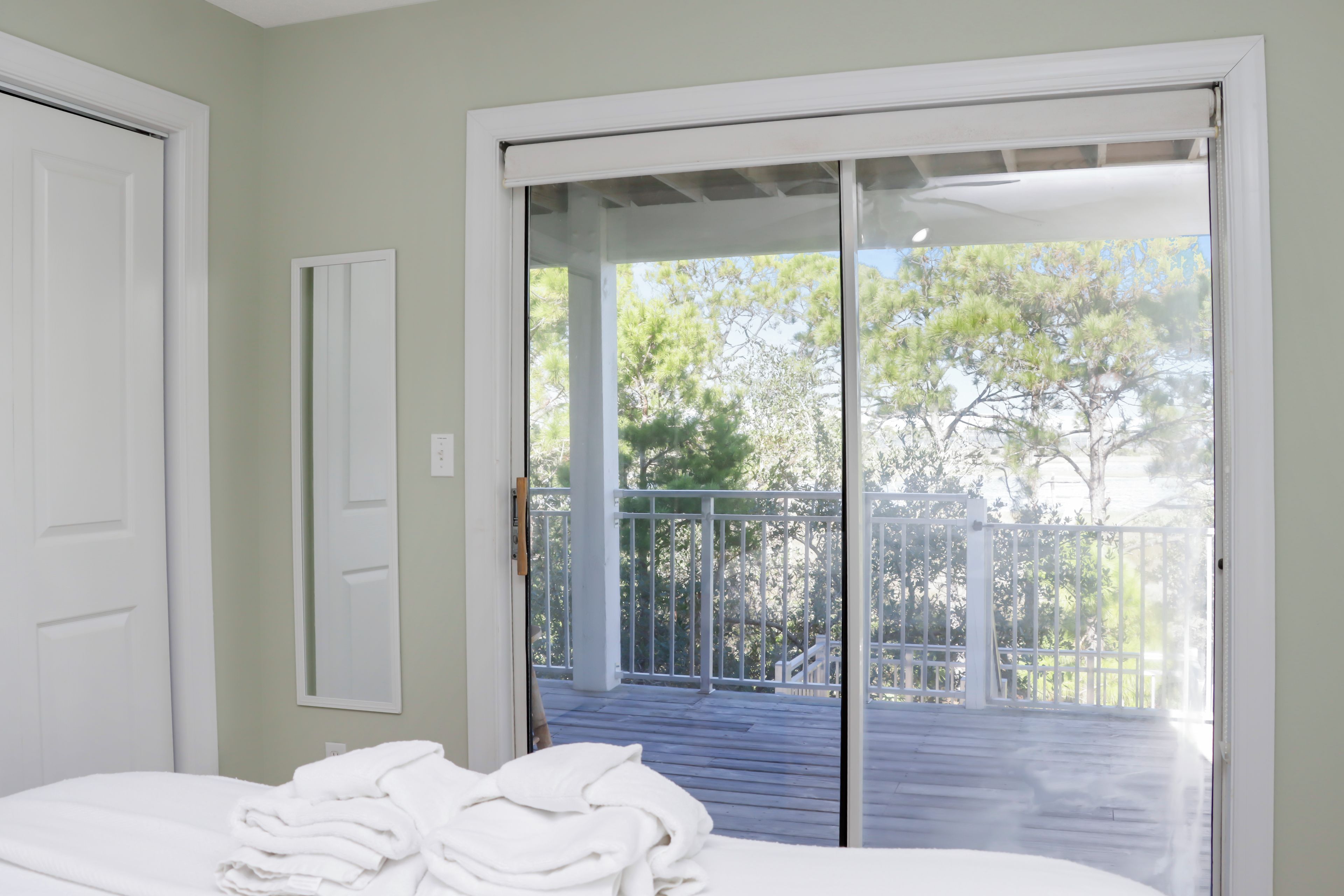 Une chambre à coucher avec un lit, une porte et une porte-fenêtre coulissante donnant sur une terrasse extérieure dotée d’une rambarde et bordée d’arbres.