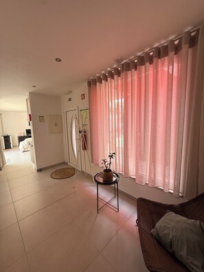 A living room with a sofa, a small table with a plant, a window with pink curtains, and a door leading to another room.
