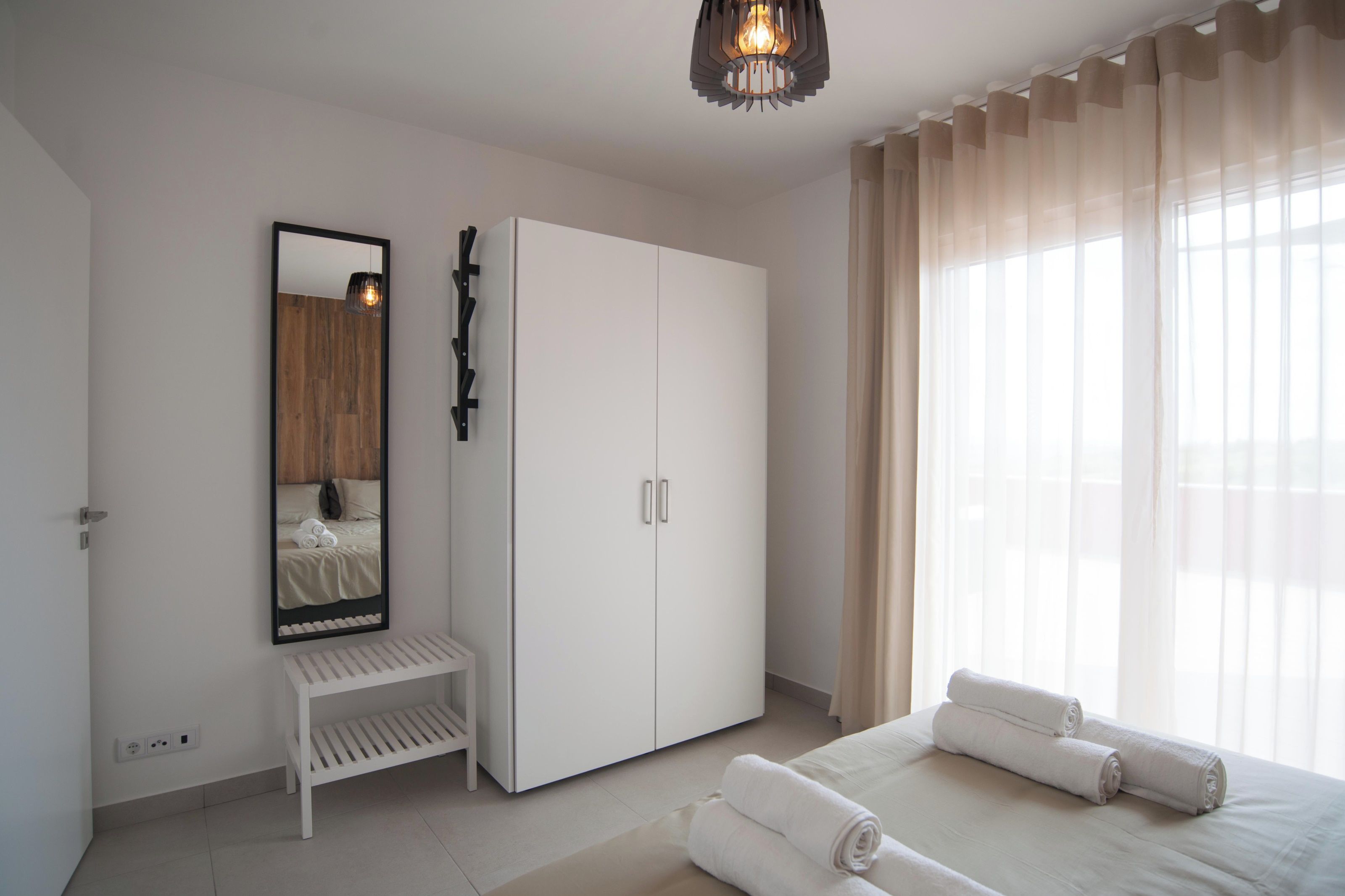 A bedroom with a bed, a wardrobe, a bench, a mirror, and a window with curtains.