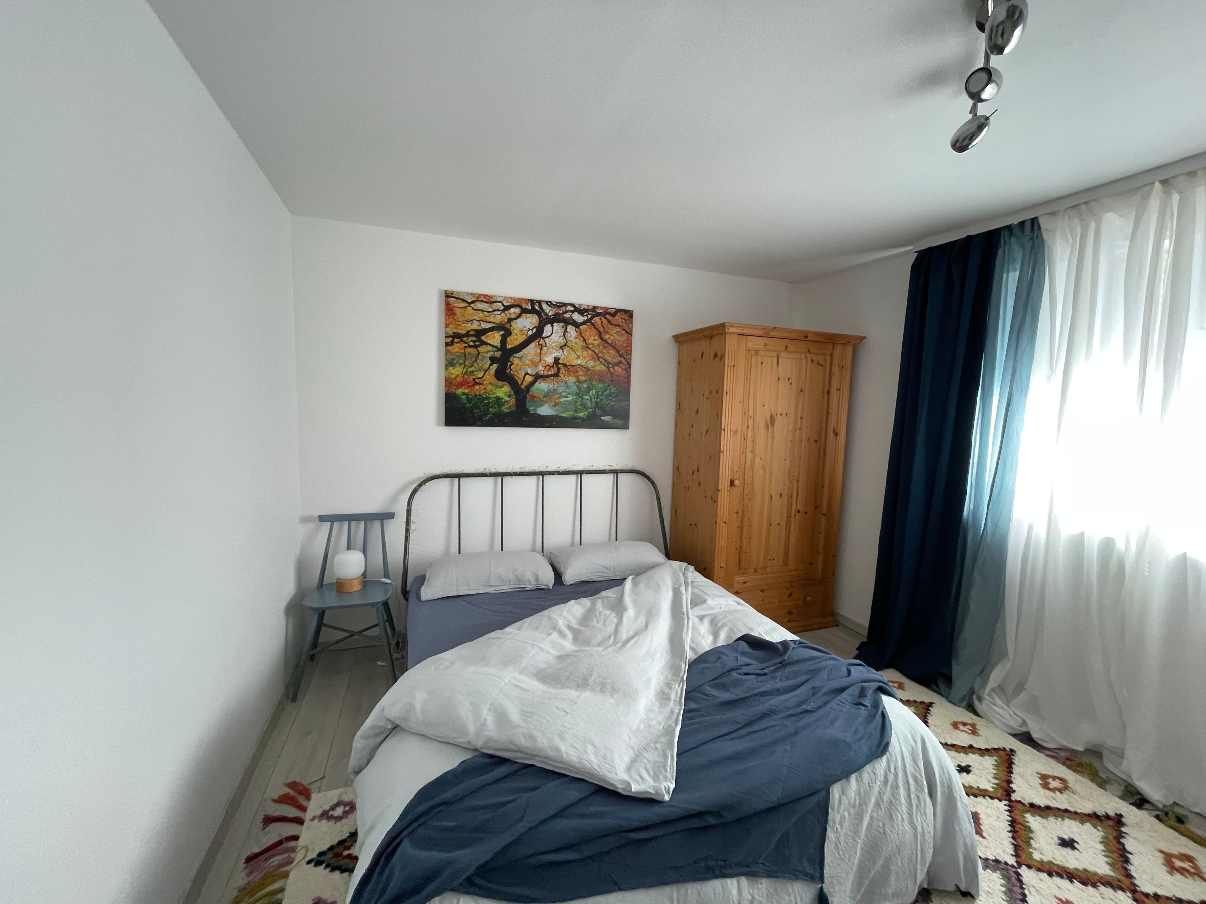 Ein Schlafzimmer mit einem Bett, einem Holzschrank, einem Bild an der Wand und einem Fenster mit Vorhängen.