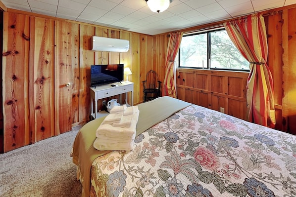 Ein Zimmer mit Holzwänden, einem Bett mit Blumenmuster auf der Tagesdecke, einem Fernseher auf einer Kommode und einem Fenster mit Vorhängen.