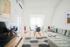 Een moderne woonkamer met een flatscreen televisie, een grijze bank en een eethoek met een glazen tafel en stoelen.