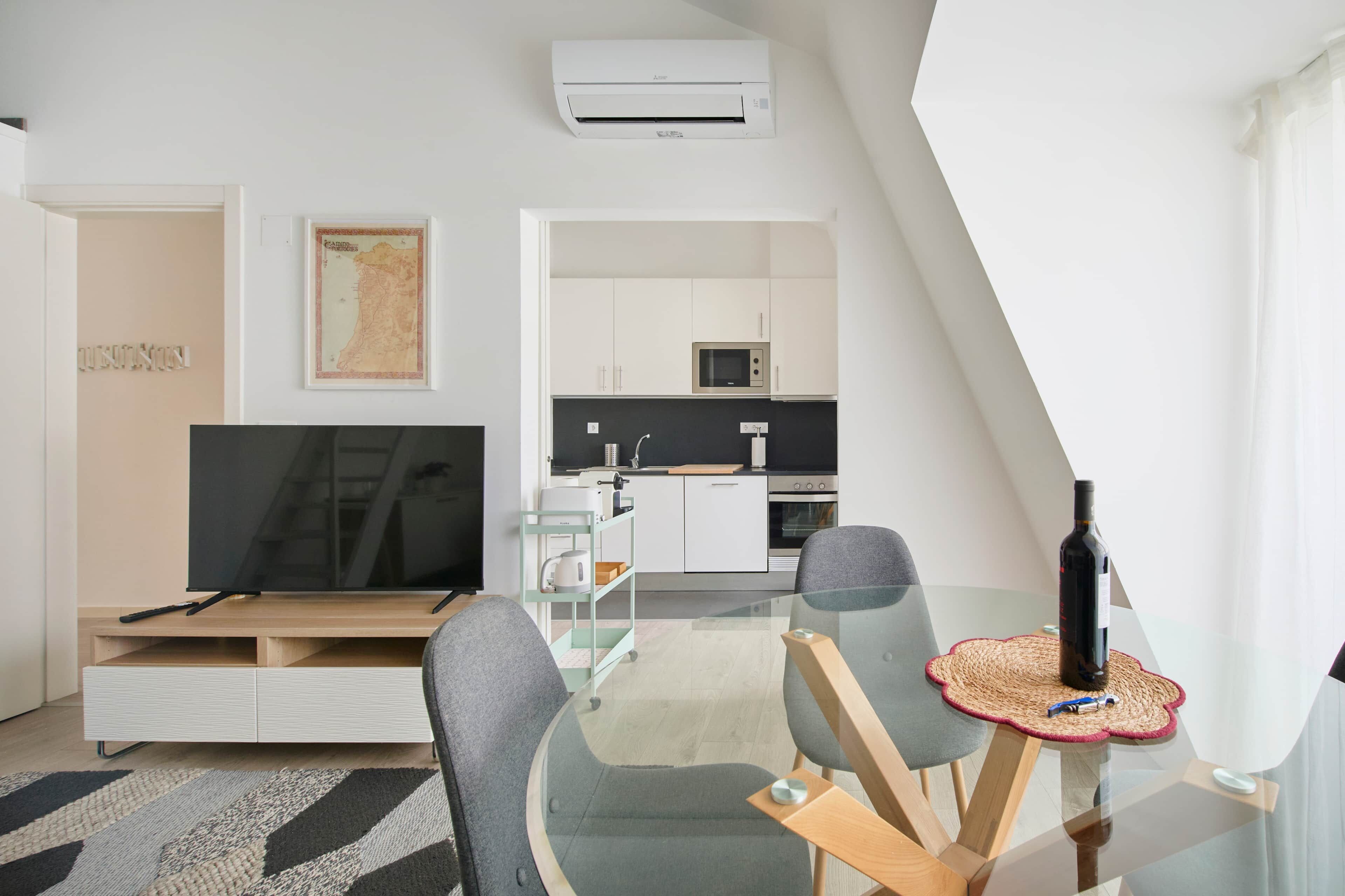 Una sala moderna con una televisión de pantalla plana, una mesa de comedor con sillas y una cocina al fondo.