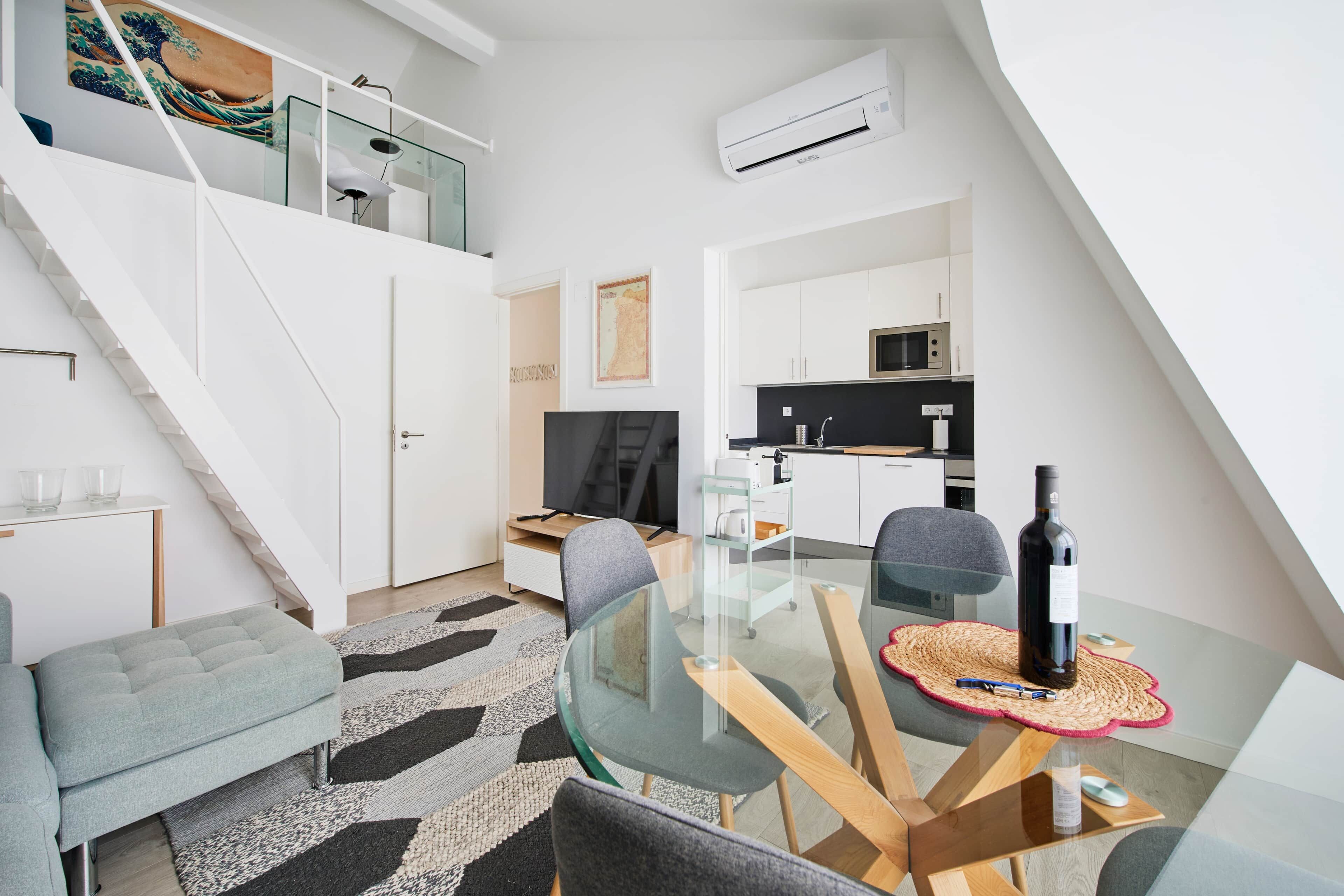 A modern apartment with a glass dining table, a sofa, a kitchen with a microwave, and a staircase.