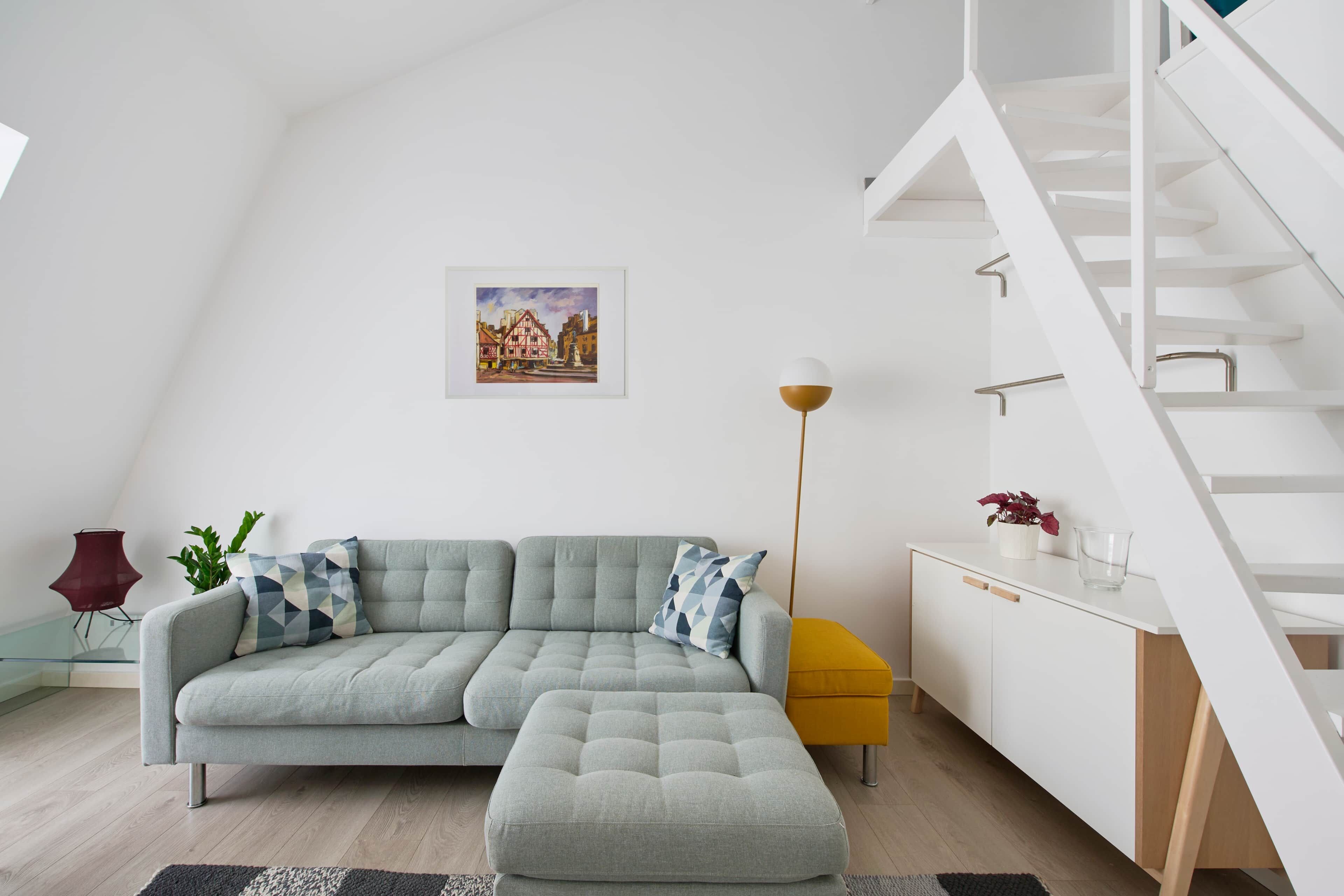 Una sala de estar moderna con un sofá modular, un reposapiés amarillo pequeño, una lámpara de pie y una escalera.