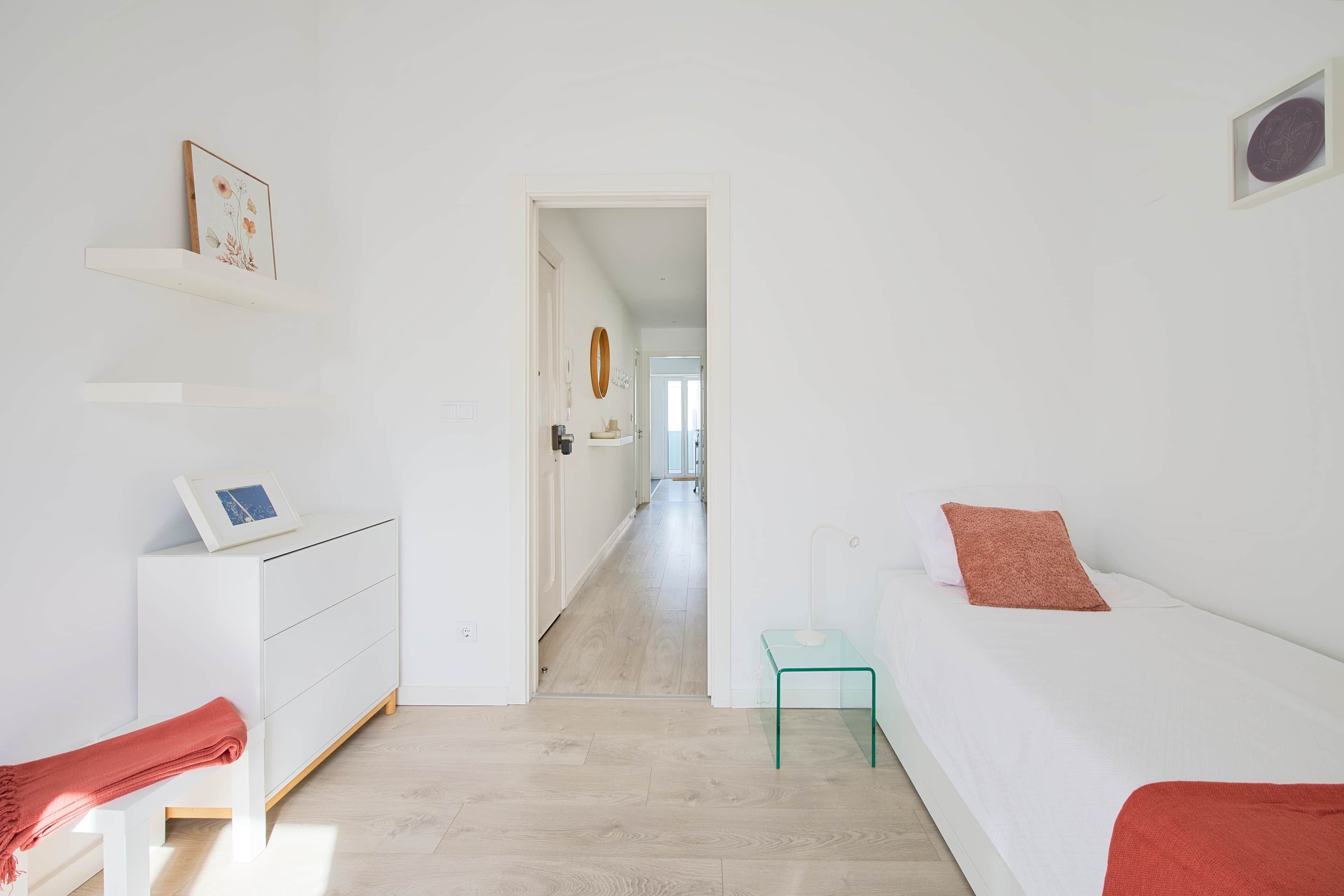 Une chambre à coucher au style minimaliste, avec un lit blanc, un petit bureau et une porte donnant sur un couloir.