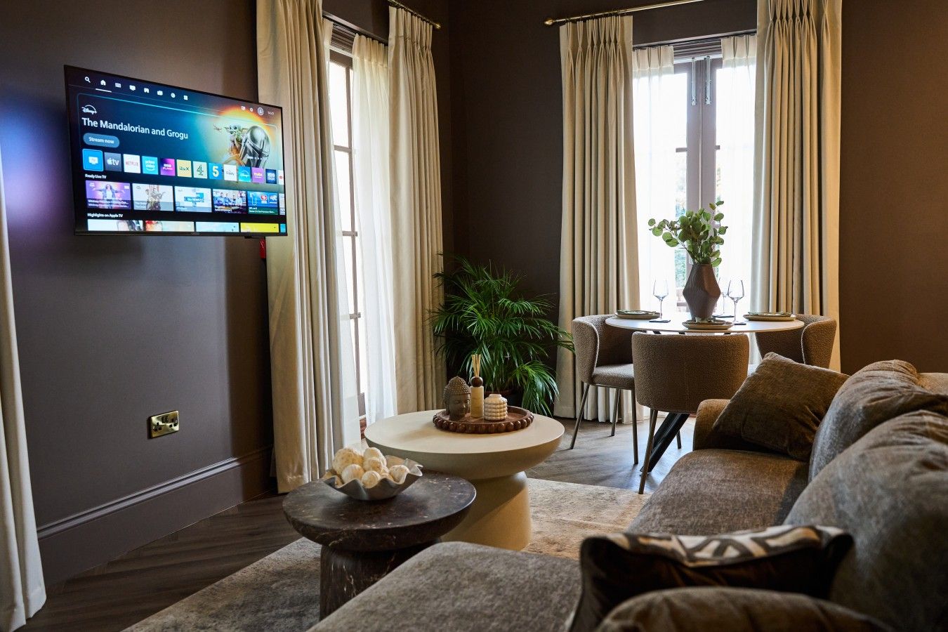 A modern living room with a flat-screen TV displaying a streaming service interface, a round coffee table with decorative items, a sofa, and a dining area with a table and chairs.