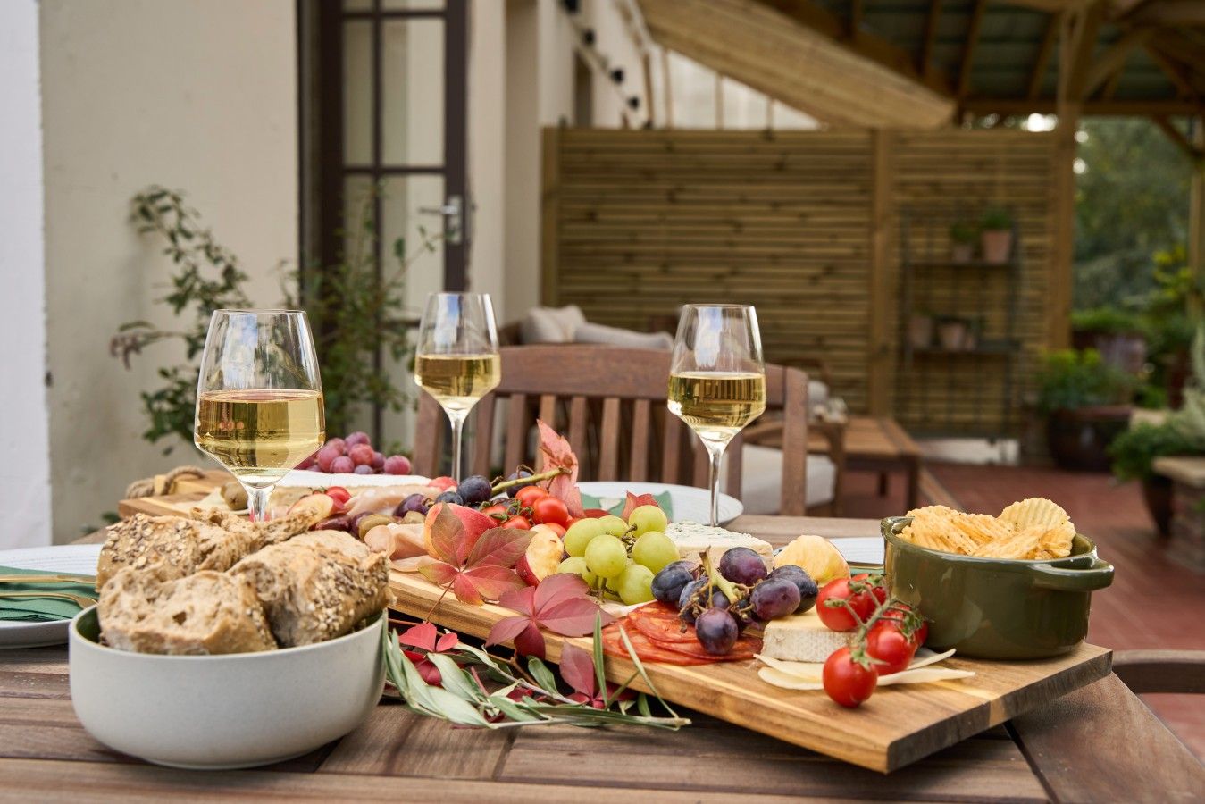 A table set with a variety of cheeses, fruits, and crackers, accompanied by glasses of white wine, in an outdoor setting with wooden furniture and plants.