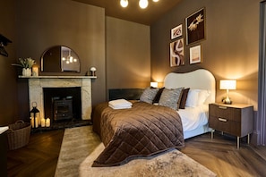 A bedroom with a fireplace, a bed with a quilted bedspread, bedside lamps, and framed artwork on the wall. - *NEW* Large Luxury Garden Terrace Suite in South Downs (Warningcamp)