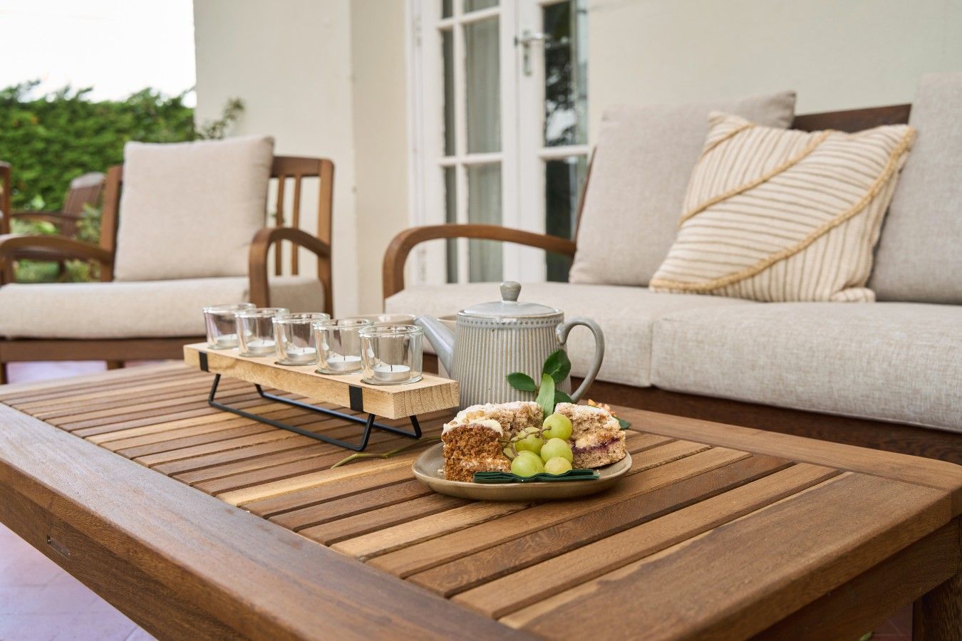 Une table en bois avec une théière et une assiette de nourriture, un plateau avec des verres, et un canapé avec des coussins en arrière-plan.