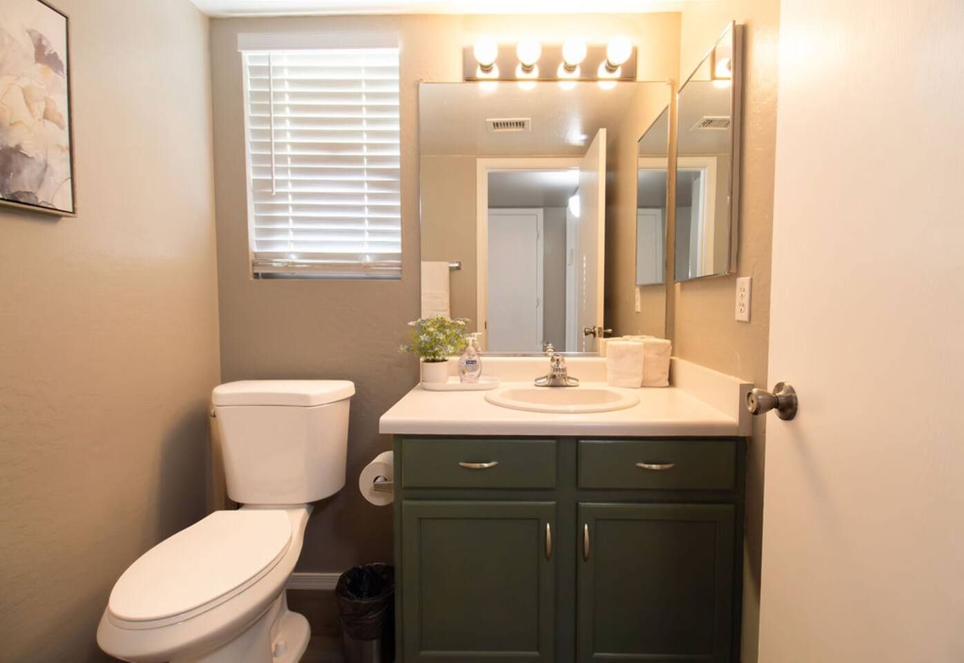 A bathroom with a toilet, sink, cabinet, mirror, and a window with blinds.