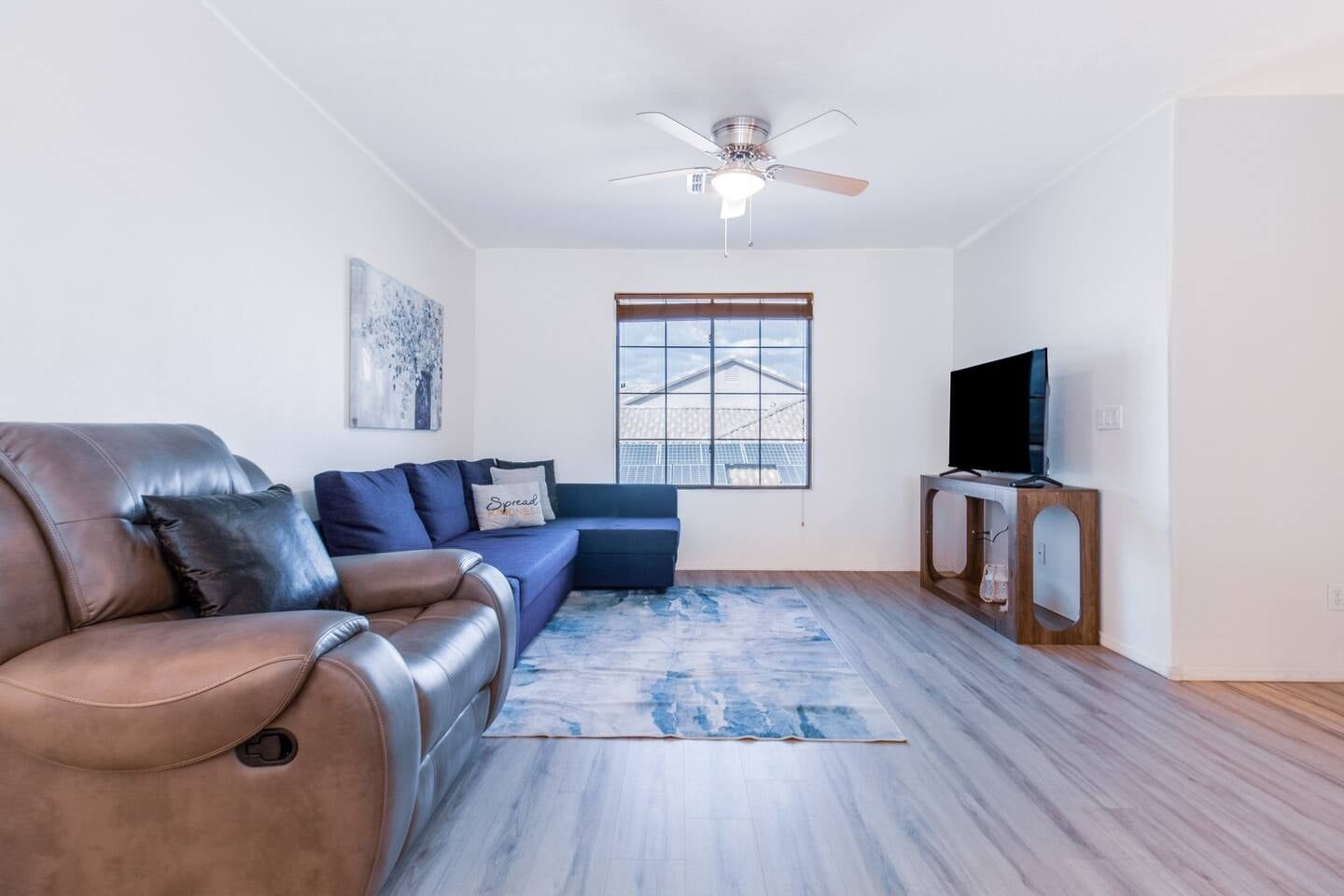 A living room with a leather sofa, a blue couch, a flat-screen TV on a wooden stand, a ceiling fan, and a window with a view of a building.