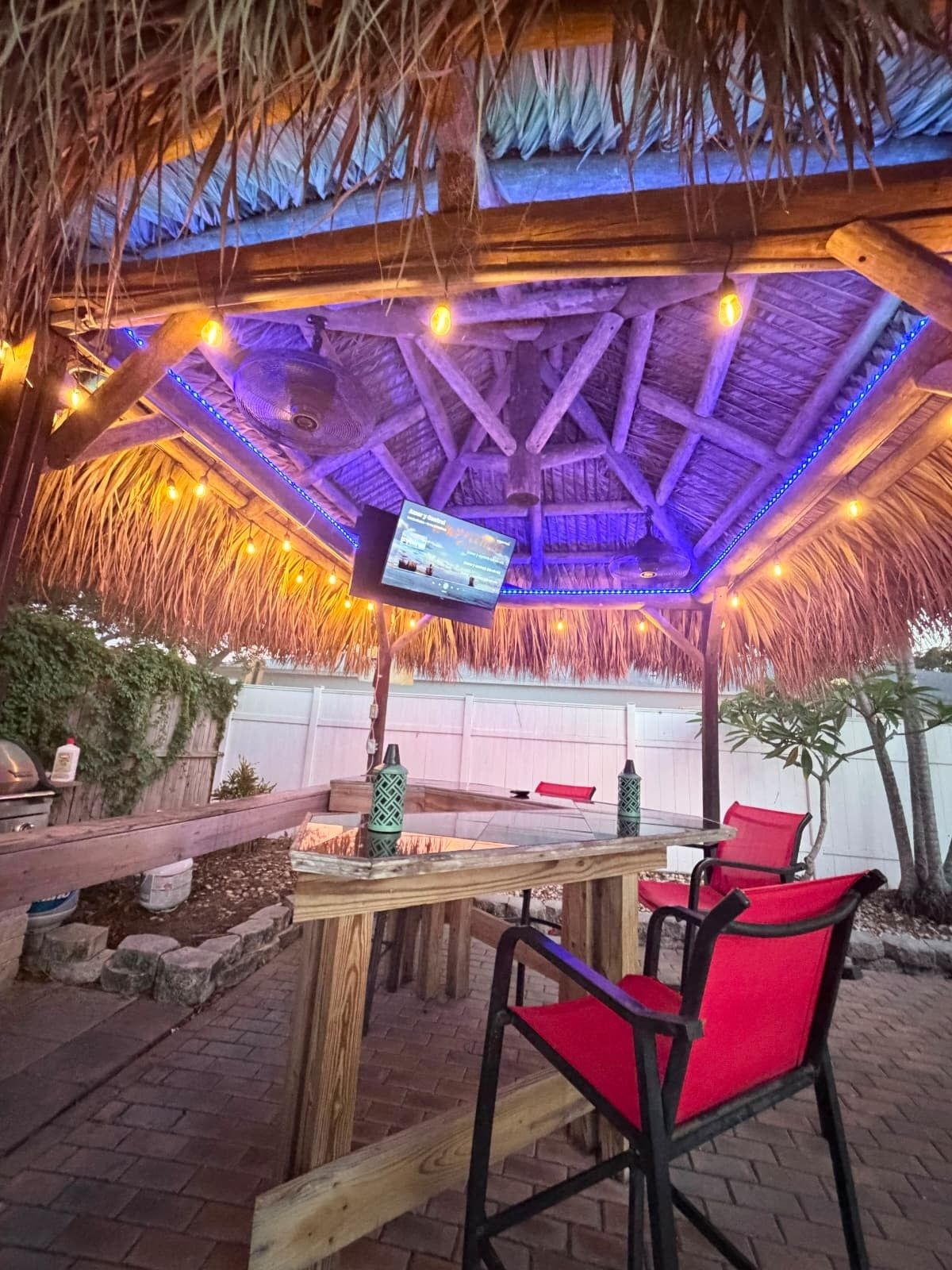 Un área de bar al aire libre con techo de paja, una pantalla de televisión y sillas rojas.