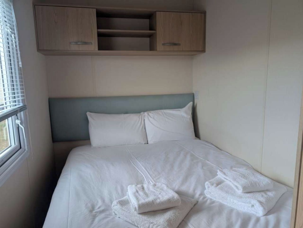 A neatly made bed with white linens and towels, a light blue headboard, and wooden cabinets above.