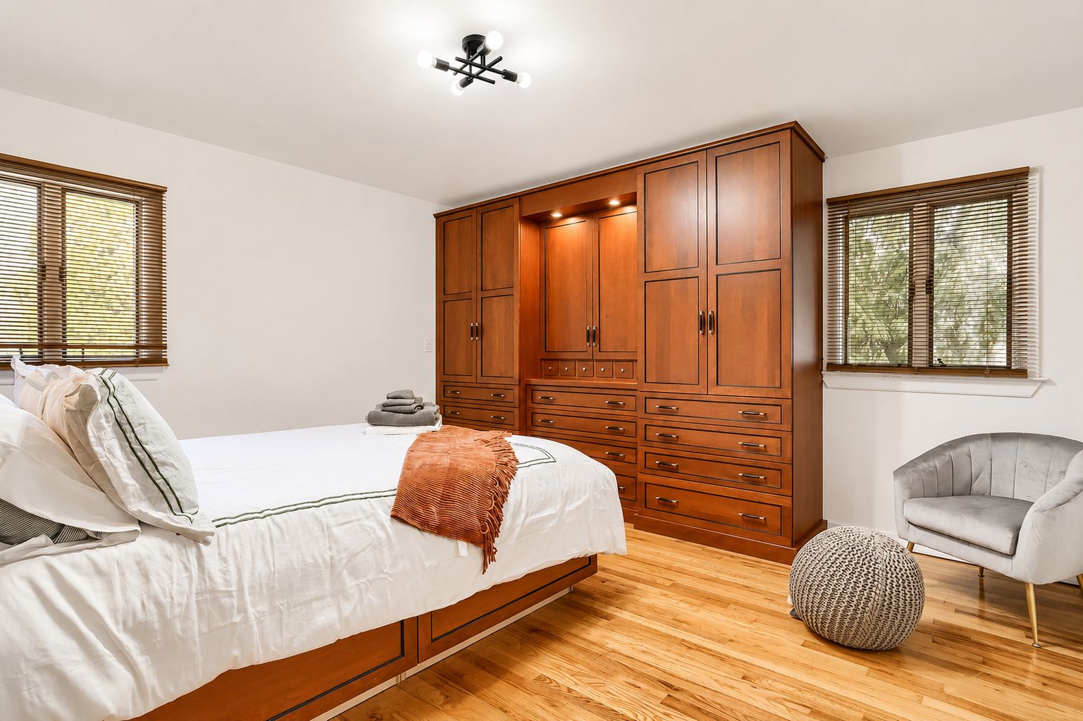 A bedroom with a large wooden wardrobe, a bed with white bedding, a grey armchair, and a round woven ottoman.