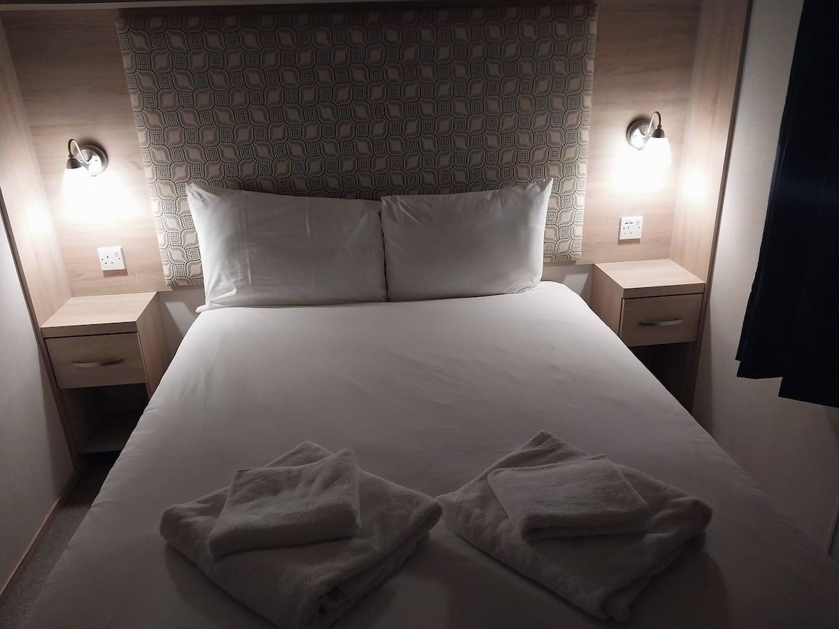 Cama bem arrumada com duas almofadas e duas toalhas dobradas sobre ela. Dois abajures estão sobre as mesas de cabeceira, um de cada lado da cama.