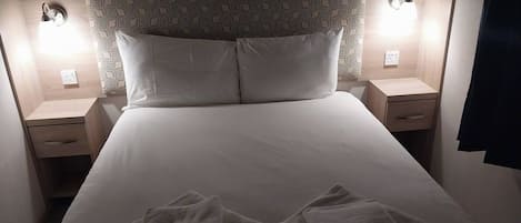 Una cama bien hecha con dos almohadas y dos toallas dobladas encima. Hay dos mesitas de noche con lámparas a cada lado de la cama.