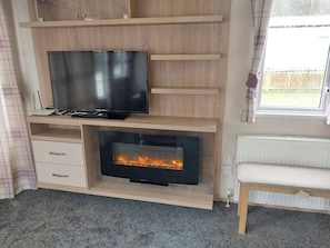 A room with a television on a wooden shelf, a fireplace below, and a bench to the right. - Bluebell corner - with small enclosed decking (Hastings)