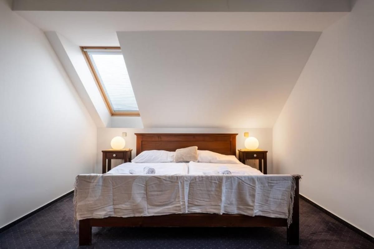 Une chambre avec un plafond en pente, un puits de lumière, un lit en bois, deux tables de chevet avec des lampes et une couvre-lit à motifs.