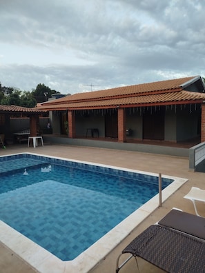 Une piscine avec une terrasse carrelée, un patio couvert doté de murs en briques et d’un toit carrelé, ainsi qu’une chaise longue.