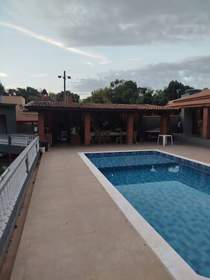 A swimming pool with a tiled deck, a covered patio area, and a view of surrounding buildings.