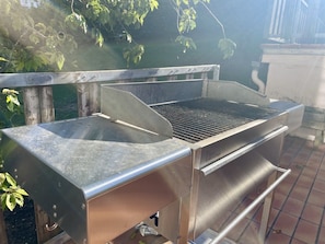 Ein Grill aus Edelstahl mit flacher Grillfläche und einer Seitenablage, draußen aufgegestellt, mit gefliestem Boden und einem Metallgeländer im Hintergrund.