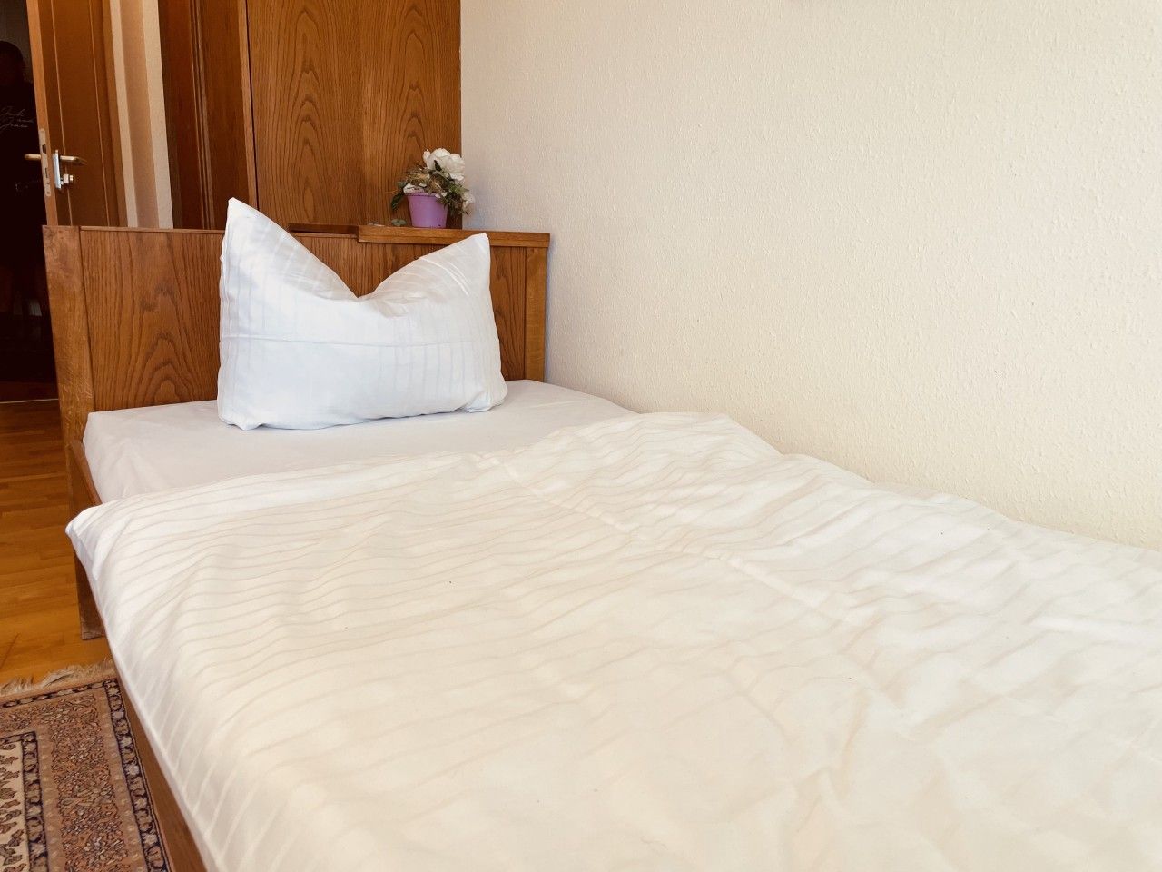 Ein Einzelbett mit weißer Matratze und Kissen, ein Holz-Kopfteil, ein kleiner Topfpflanze und ein Holzschrank.