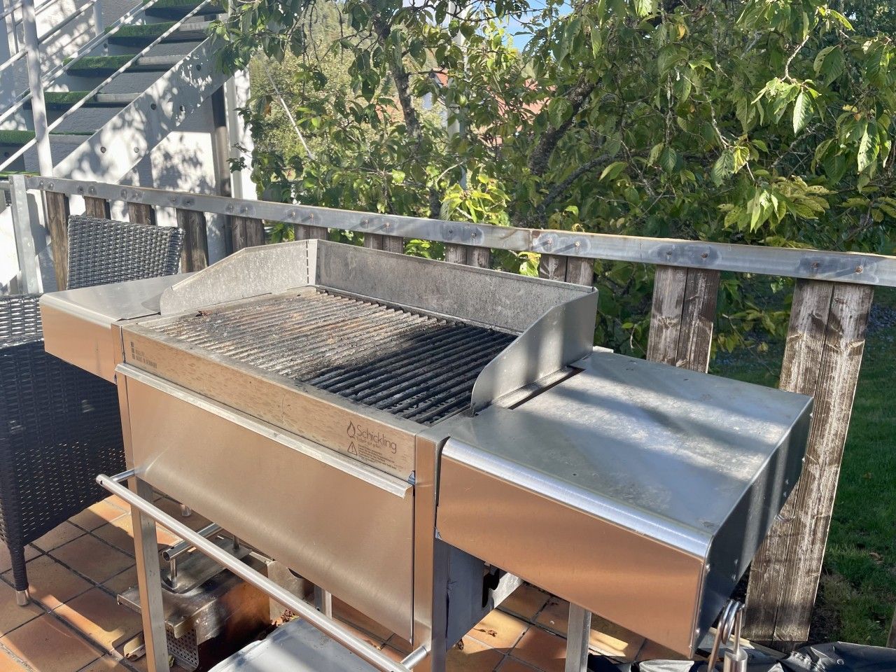A stainless steel outdoor grill with a built-in smoker, placed on a patio with a wooden fence and greenery in the background.