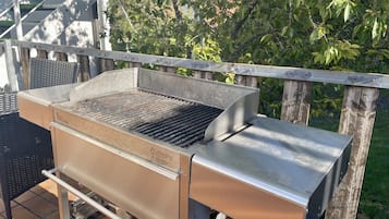 A stainless steel outdoor grill with a built-in smoker, placed on a patio with a wooden fence and greenery in the background.