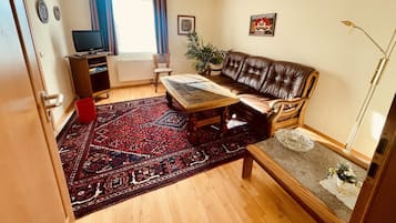 Una sala de estar con un sofá, una mesa de centro, un televisor, un tapete rojo y una planta.