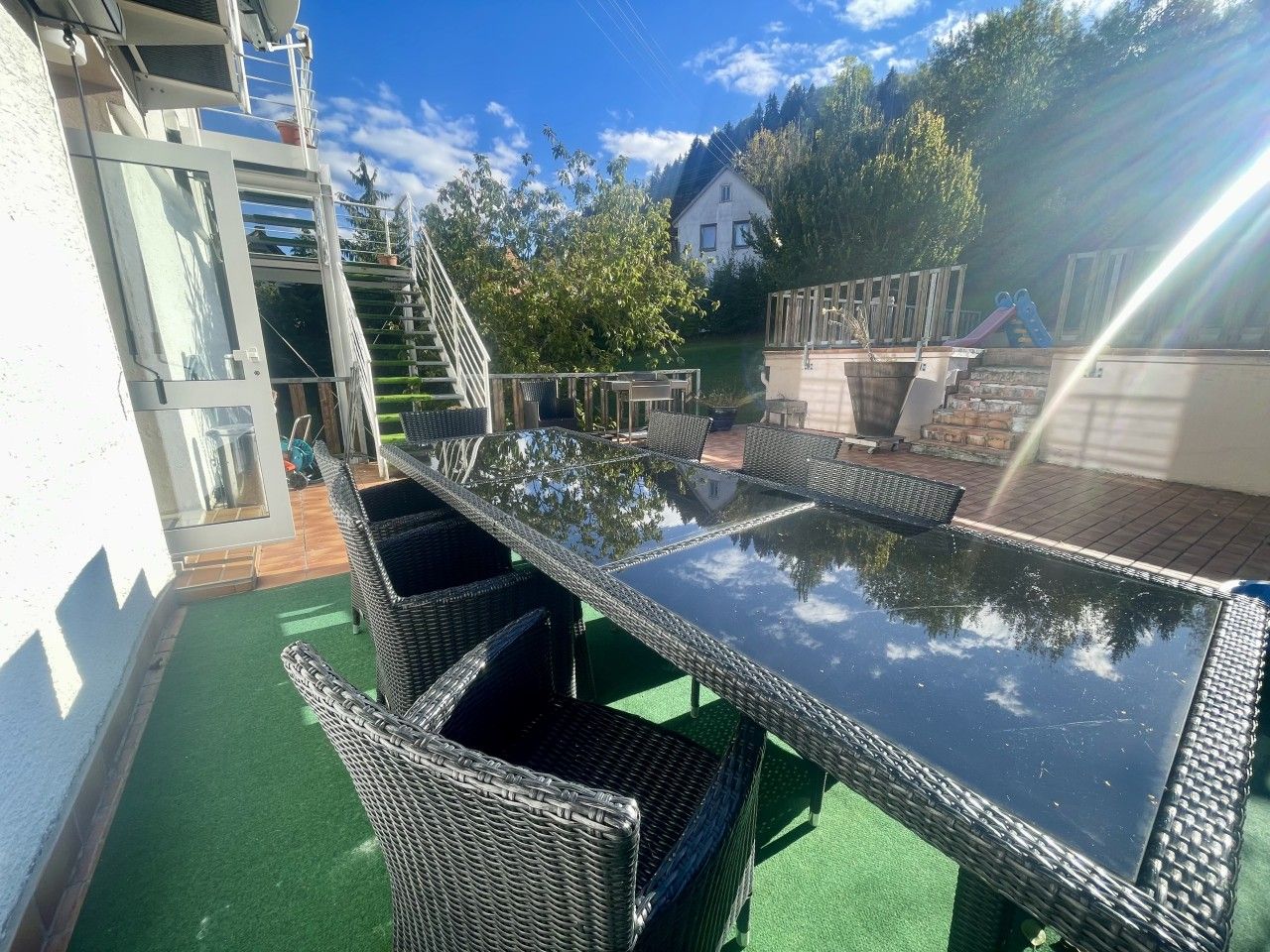 Une terrasse extérieure avec une table à plateau de verre et des chaises en osier, entourée d’une pelouse artificielle verte et d’un bâtiment blanc en arrière-plan.