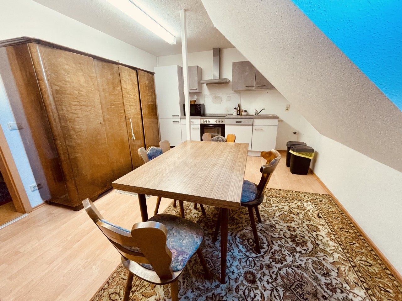 Un coin repas avec une table et des chaises en bois, un tapis à motifs et une cuisine en arrière-plan.