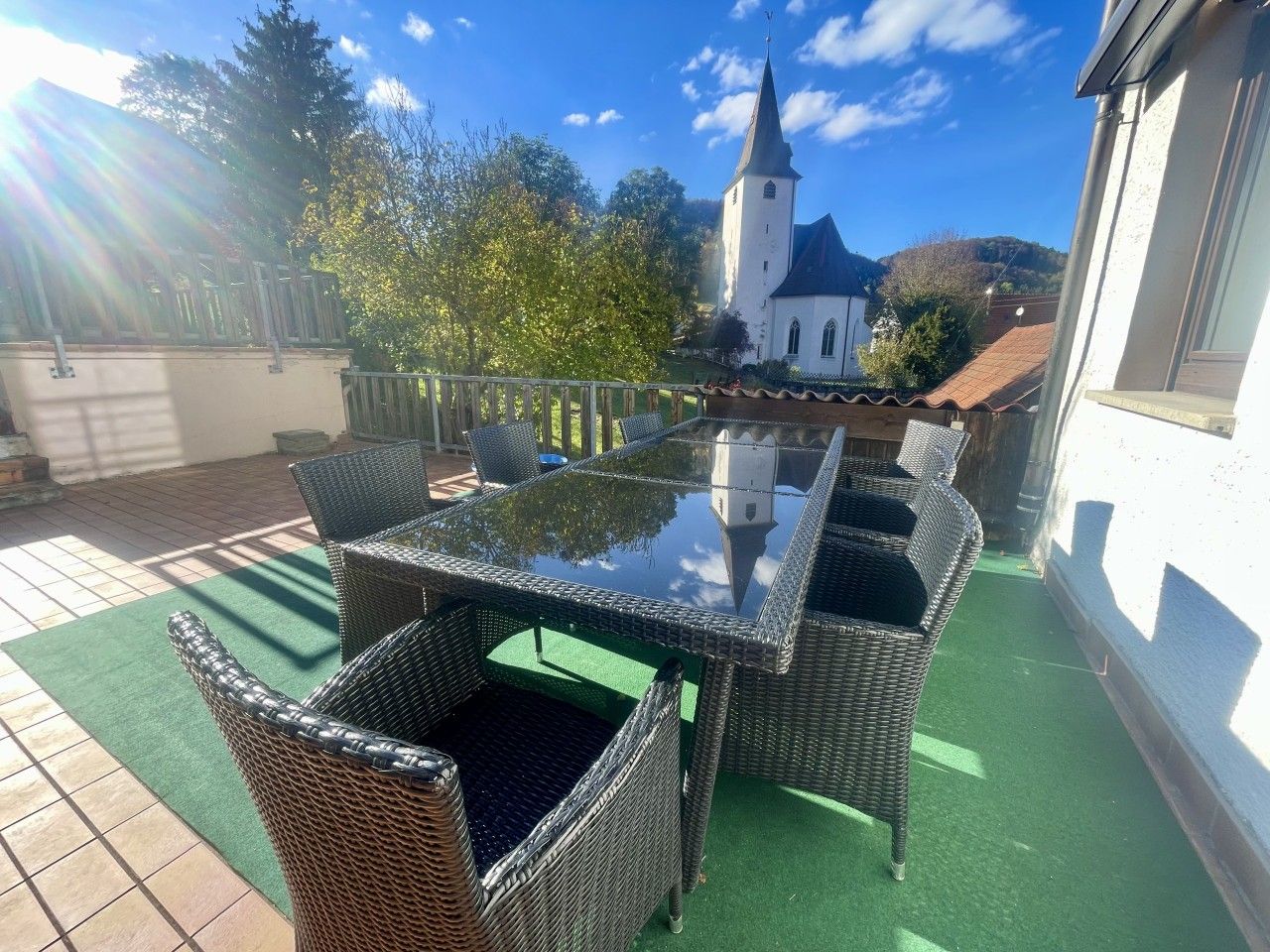 Une terrasse extérieure avec des meubles en osier et une table en verre, une vue sur une église et un espace recouvert d’un gazon synthétique vert.