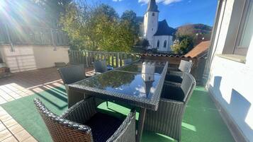 Une terrasse extérieure avec des meubles en osier et une table en verre, une vue sur une église et un espace recouvert d’un gazon synthétique vert.