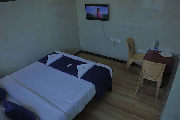 A hotel room with a bed, a desk, a chair, and a television mounted on the wall. - Anugraha Lodge (Ramanagara)