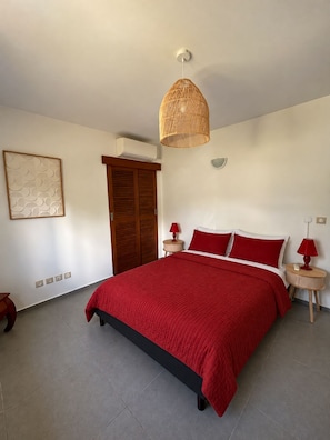 Quarto com cama, luminária pendente, ar-condicionado fixado na parede e uma pequena mesa de cabeceira com abajur.