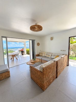 Ein modernes Wohnzimmer mit einem großen Rattan-Sofa, einem Esstisch und Blick auf den Ozean.