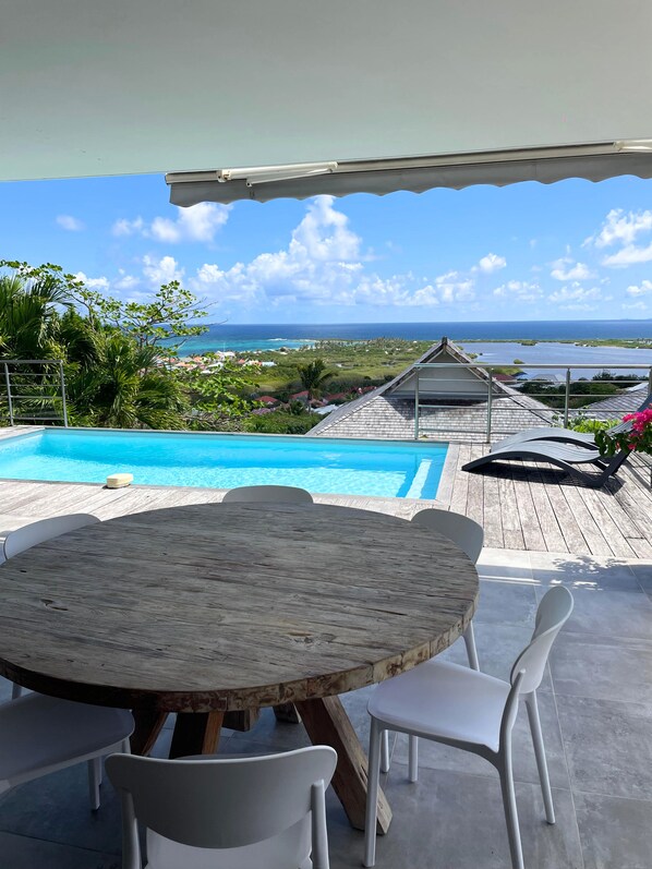 Um deck de madeira com piscina, cadeiras brancas e uma mesa, com vista para a praia e o oceano.