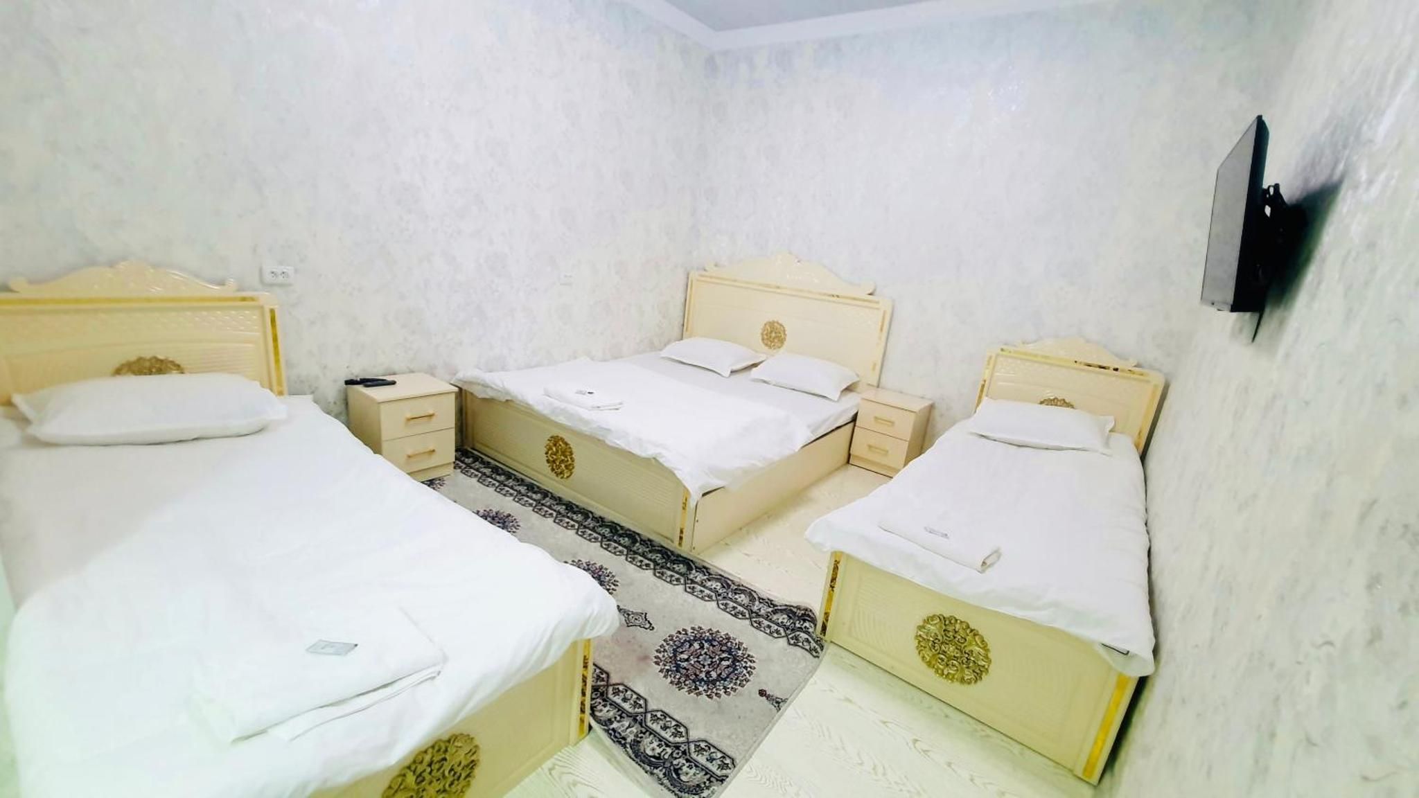 A room with three beds, each with a white mattress and matching pillows, arranged in a corner. There is a small bedside table with a lamp on one side and a drawer on the other. The walls are plain, and there is a patterned rug on the floor.