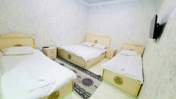 A room with three beds, each with a white mattress and matching pillows, arranged in a corner. There is a small bedside table with a lamp on one side and a drawer on the other. The walls are plain, and there is a patterned rug on the floor.